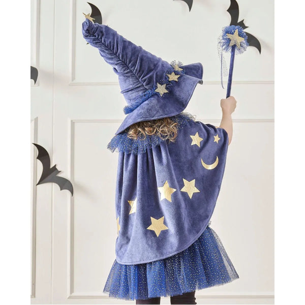 Child in a blue wizard costume with star patterns, holding a wand against a white wall with black bat decorations.