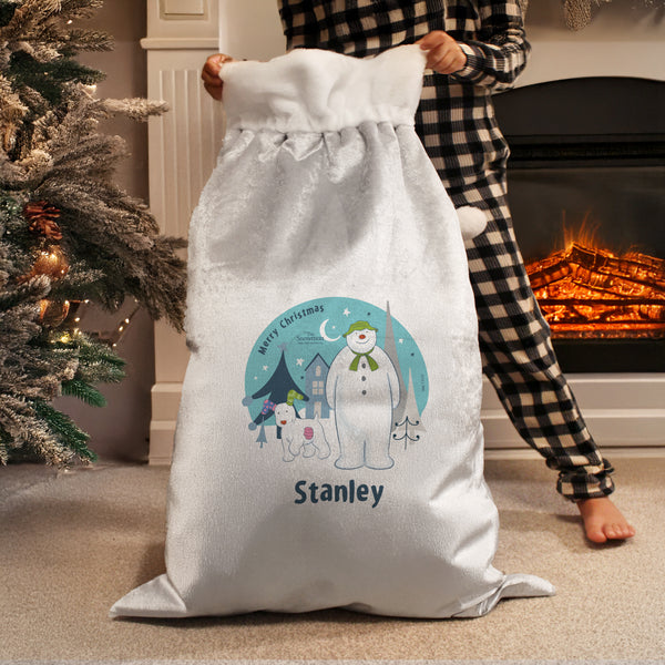White Christmas sack with a snowman design and 'Stanley' text, held by a person in checkered pajama pants in front of a fireplace.
